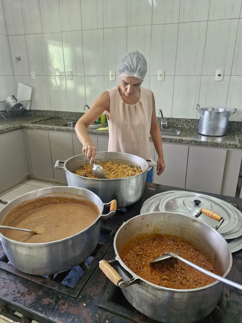 Foto ilustrativa: mulher de touca preparando arroz, feijão e molho em grandes panelas, em uma cozinha comunitária